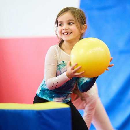 Petite fille souriante faisant de la gym avec un ballon jaune lors d'un stage Toup’ti Gym, développant ses capacités motrices et sa confiance en elle